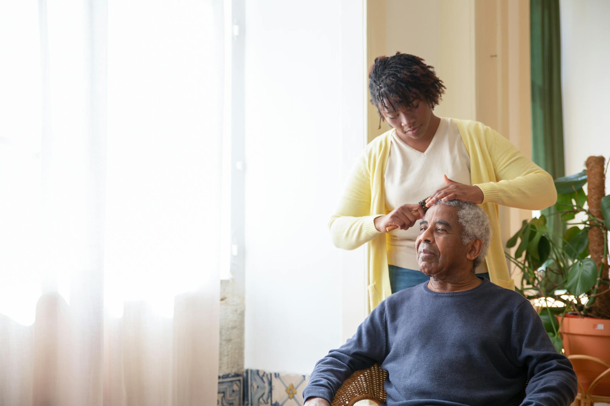 A caregiver combing an elderly man's hair near a sunlit window indoors.